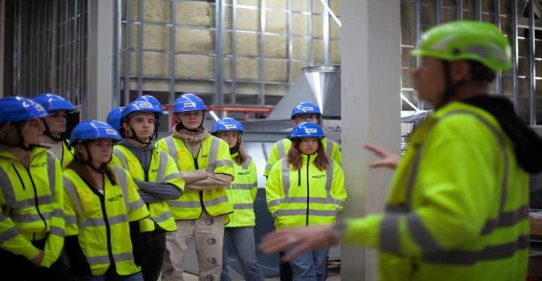 Åtta elever i varselvästar som står och lyssnar på en manlig byggledare.