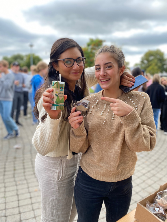 två elever som tittar in i kameran och håller upp fika.